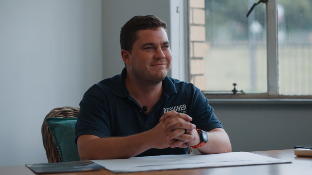 designer pool covers A man in a designer polo shirt sits at a table, smiling slightly, with his hands clasped together in front of him, in a room with a window.